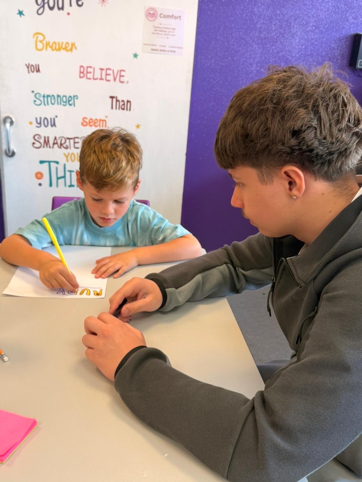 An older young person helping a younger child colour in the letters of a name design